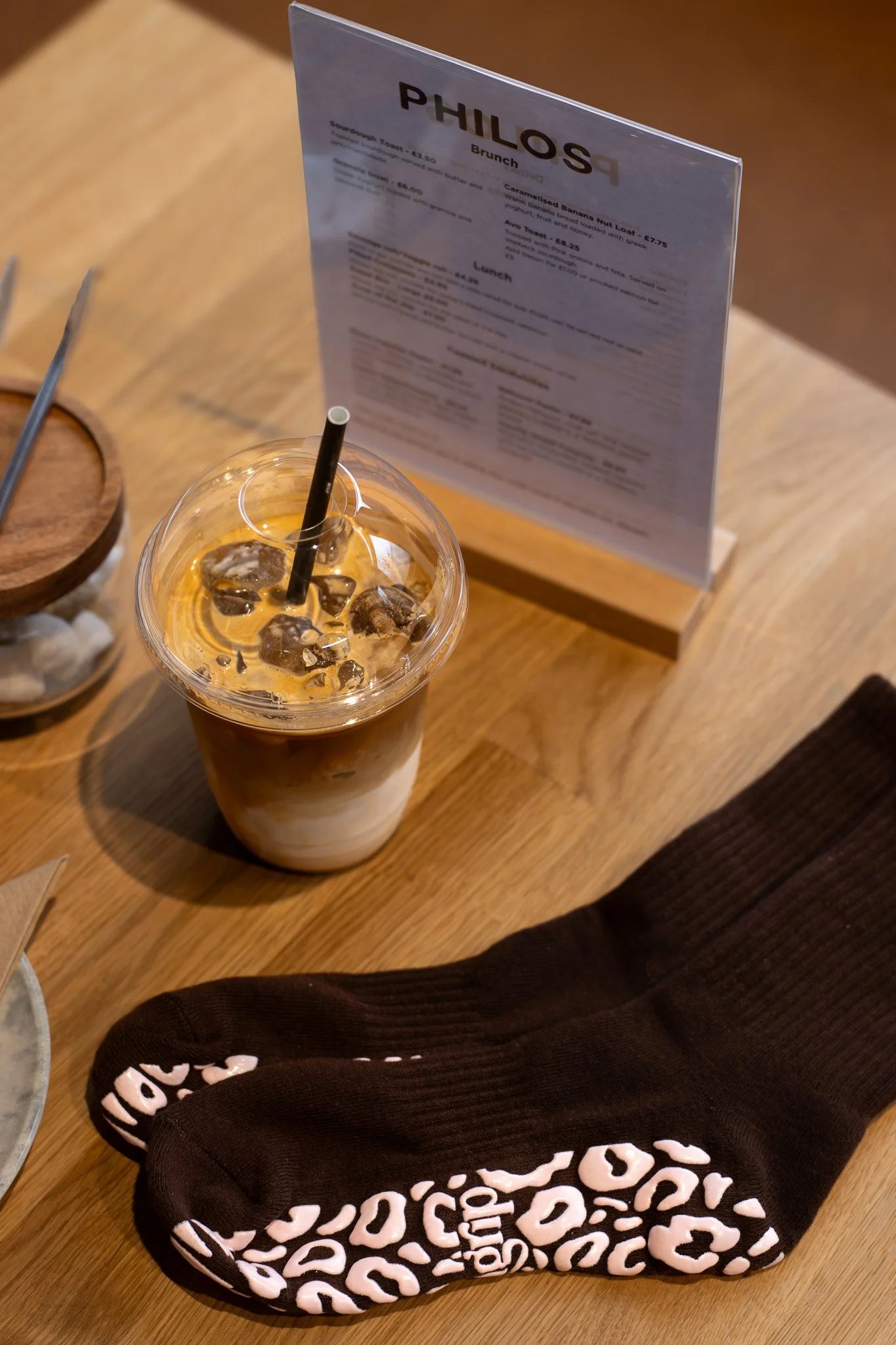 A plastic cup of iced coffee on a wooden table in front of a menu stand, with a leopard print hoodie partially visible in the lower part of the image. GetAGrip Apparel.