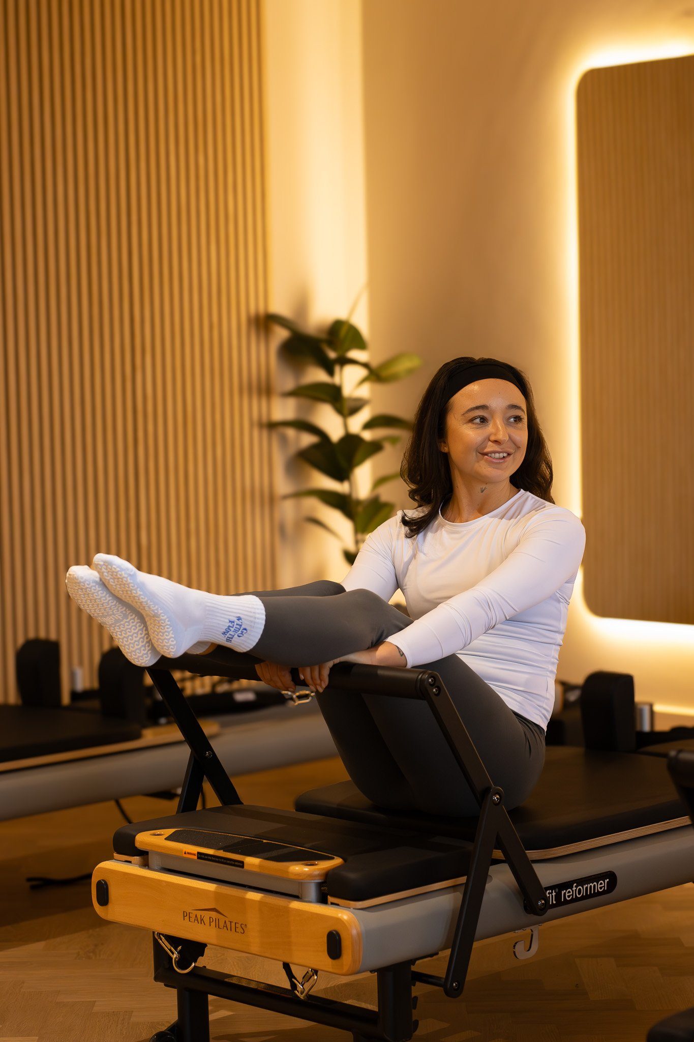 A woman practicing Pilates on a reformer machine in a modern studio with wooden walls and a potted plant in the background. GetAGrip Apparel.