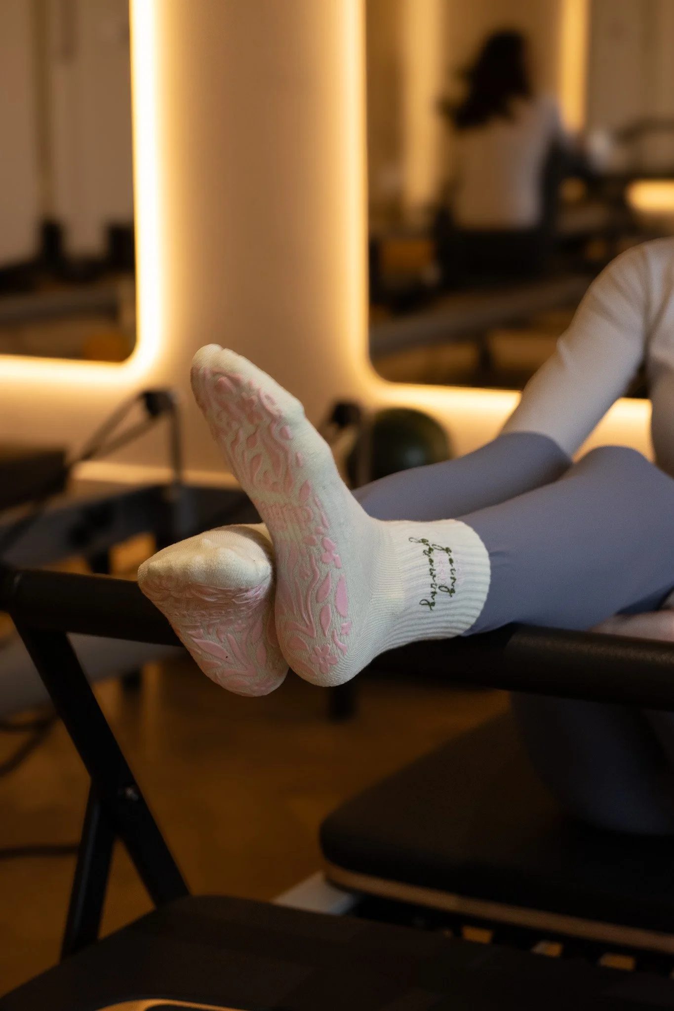 Close-up of a person's foot wearing a white medical sock with pink patterns, resting on a black chair in a room with warm lighting. GetAGrip Apparel.