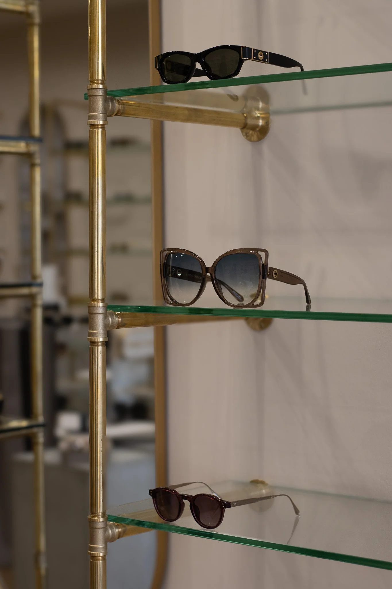 Three pairs of sunglasses on a glass and brass display shelf, with the top pair being black and the bottom pair brown, and a faded background. Linda Farrow.