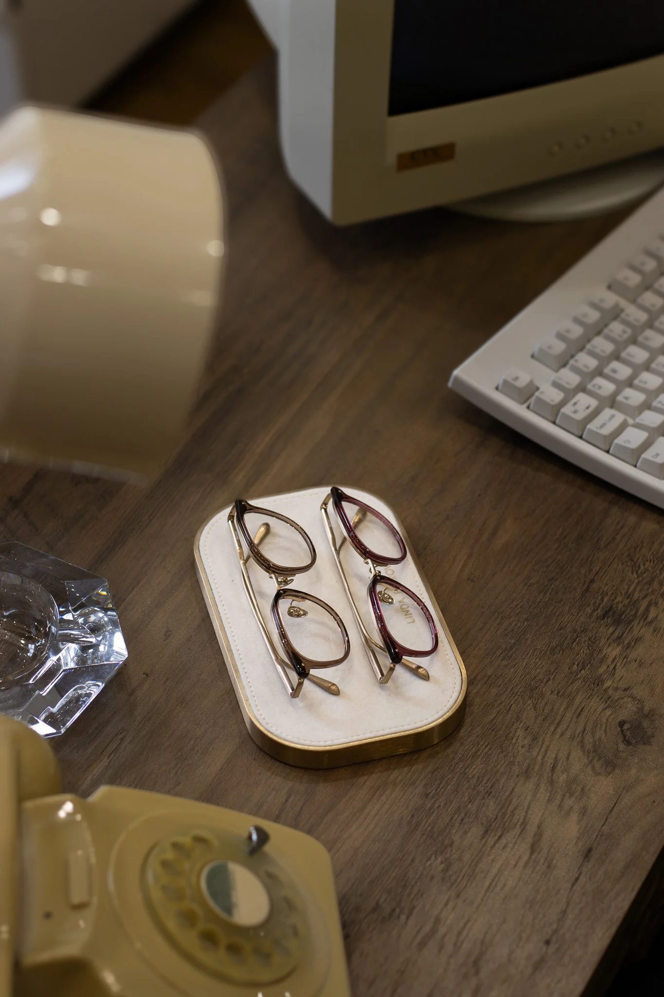 A pair of glasses resting on a white and gold case on a wooden surface with a vintage computer monitor and keyboard in the background. Linda Farrow.