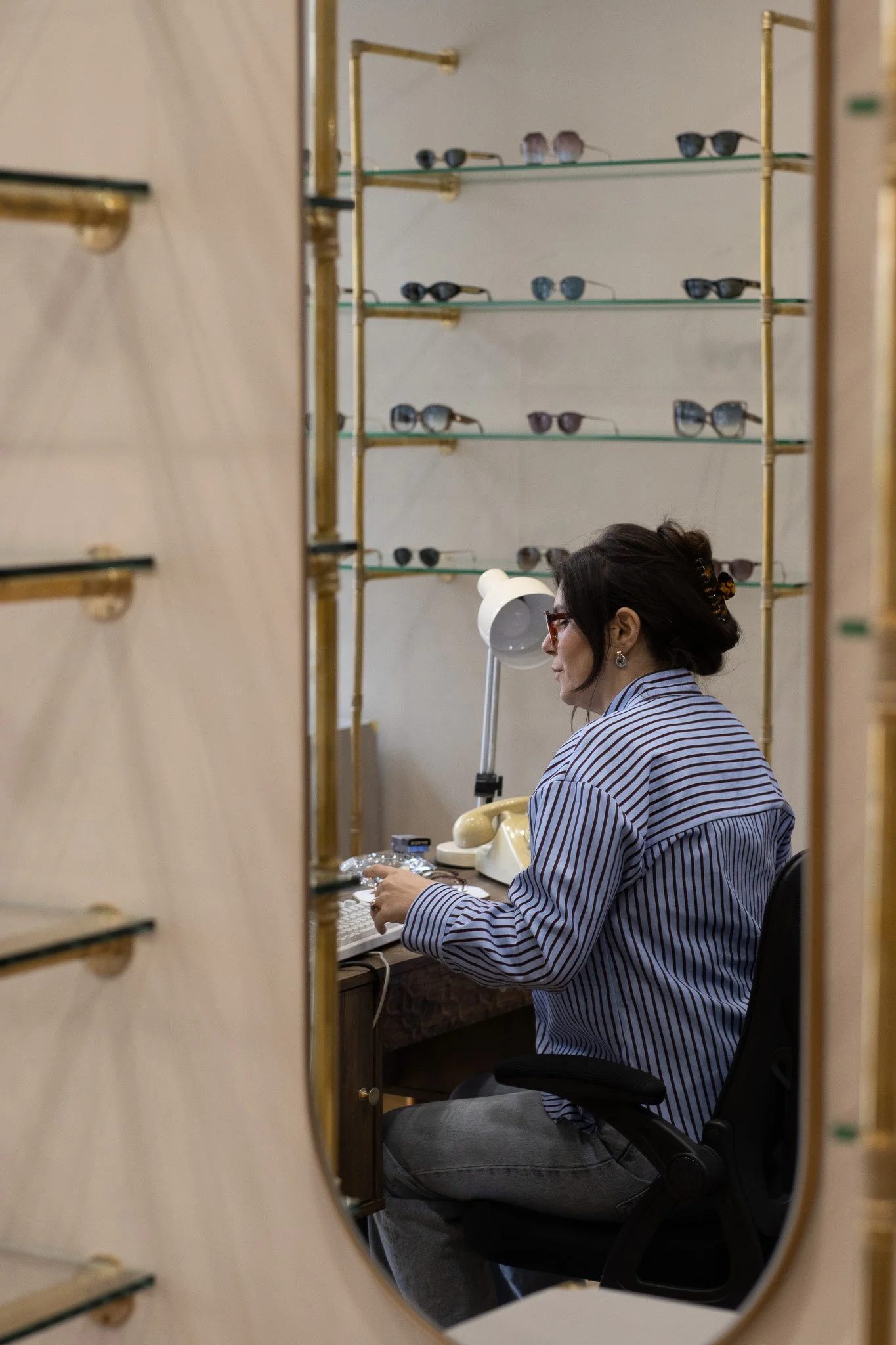 A woman with dark hair and glasses, sitting at a desk in front of a mirror, working on a laptop. The image is reflected in the mirror, showing multiple pairs of sunglasses on shelves behind her. Linda Farrow.
