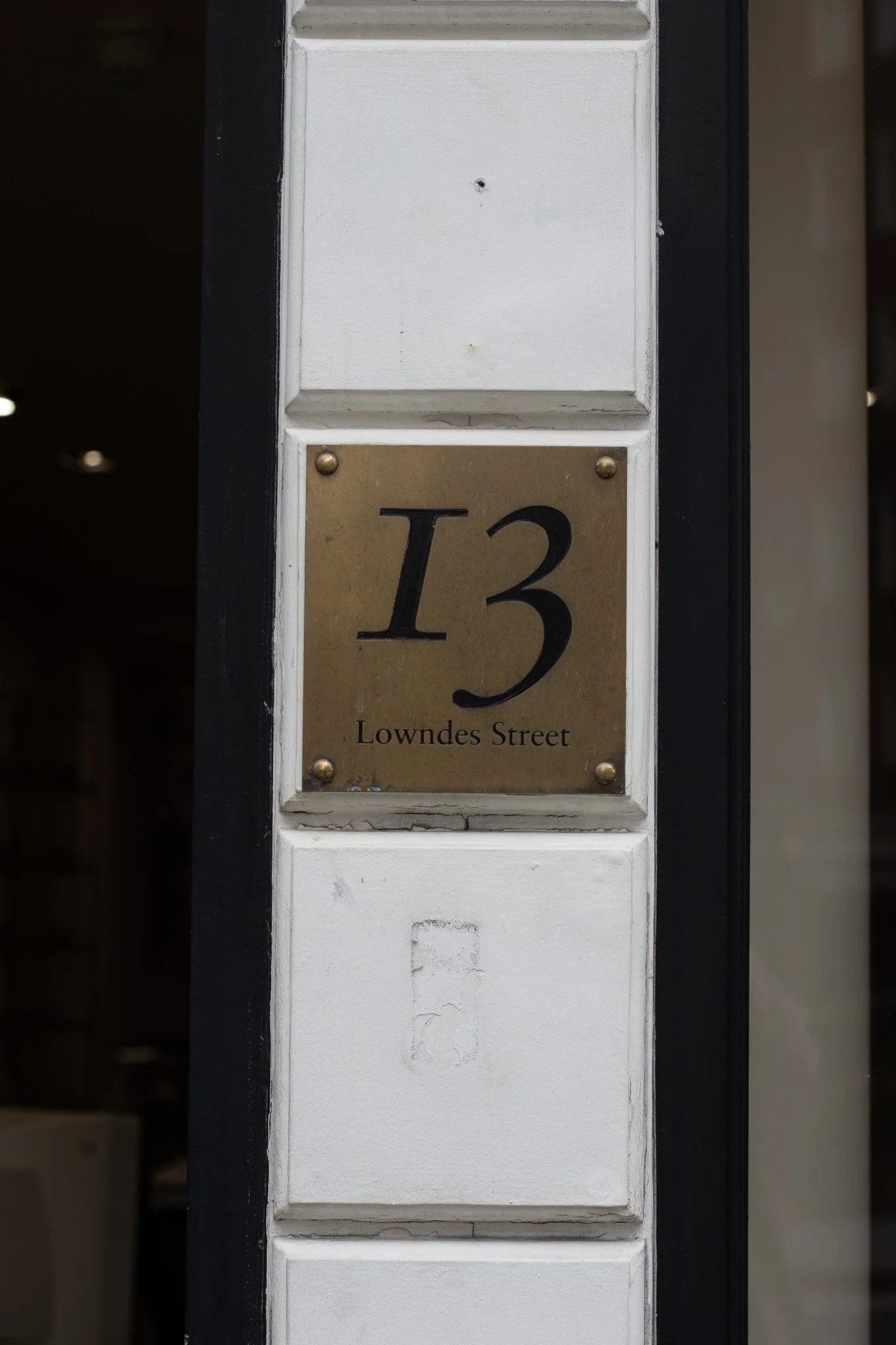 A house number plaque displaying '13 Lowndes Street' mounted on a white post with a black border; the plaque has a gold-colored background with black lettering. Linda Farrow.