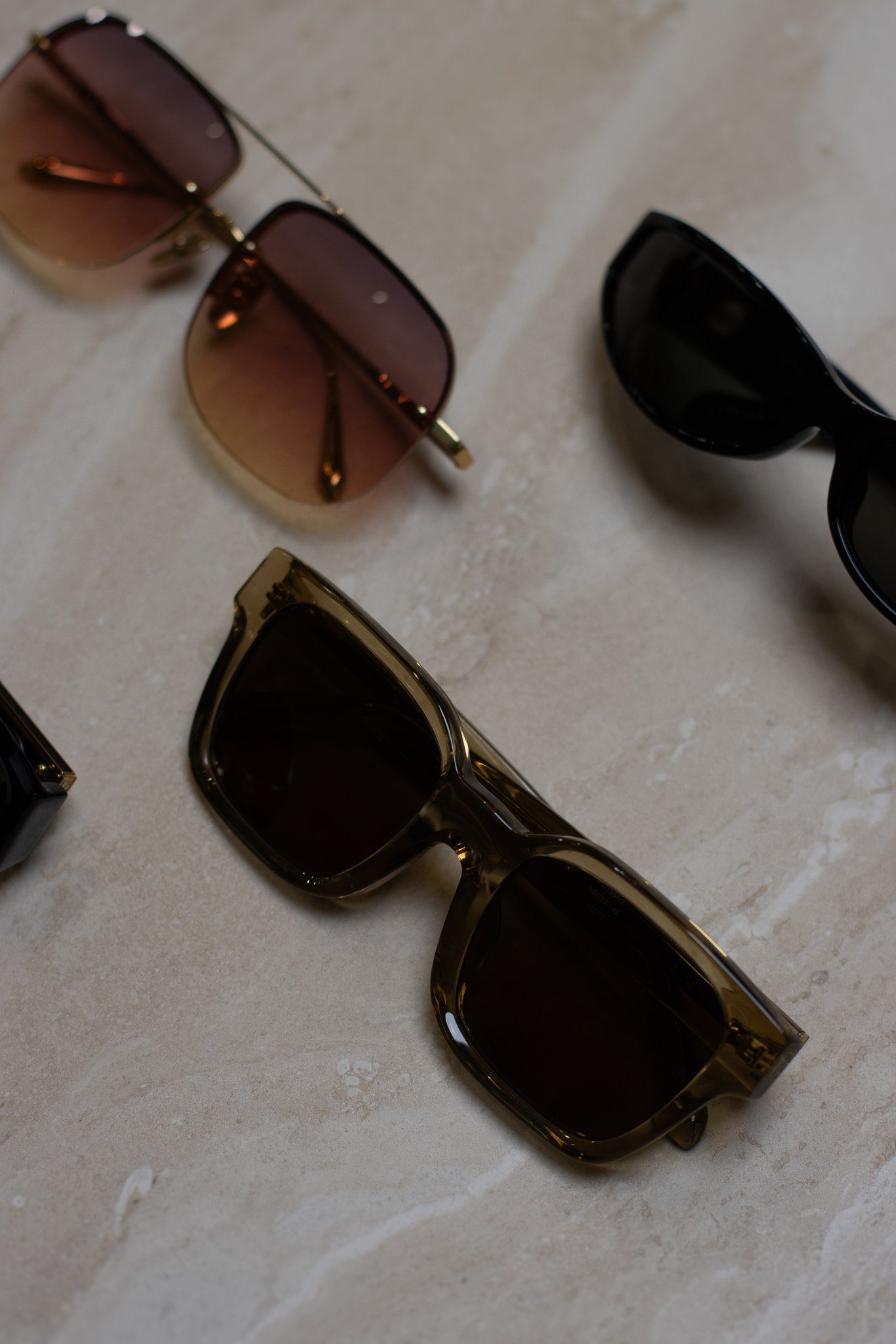 A collection of sunglasses with various frame styles and tints, including gold, black, and tortoise shell, arranged on a light-colored stone surface. Linda Farrow.