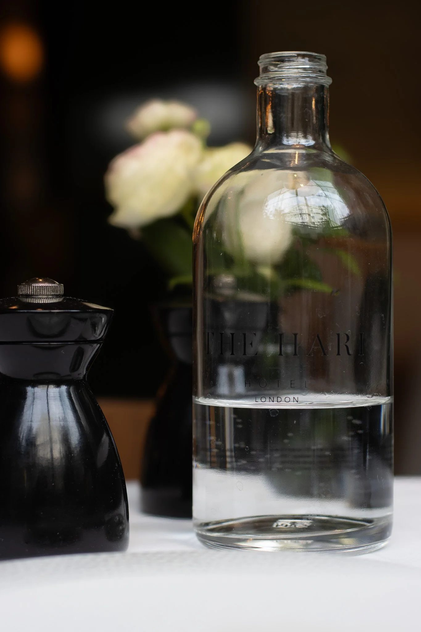 A clear glass bottle with the words 'The Hari Hotel London' etched on it, surrounded by black ceramic containers with silver lids, and blurred flowers in the background. The Hari, Il Pampero.