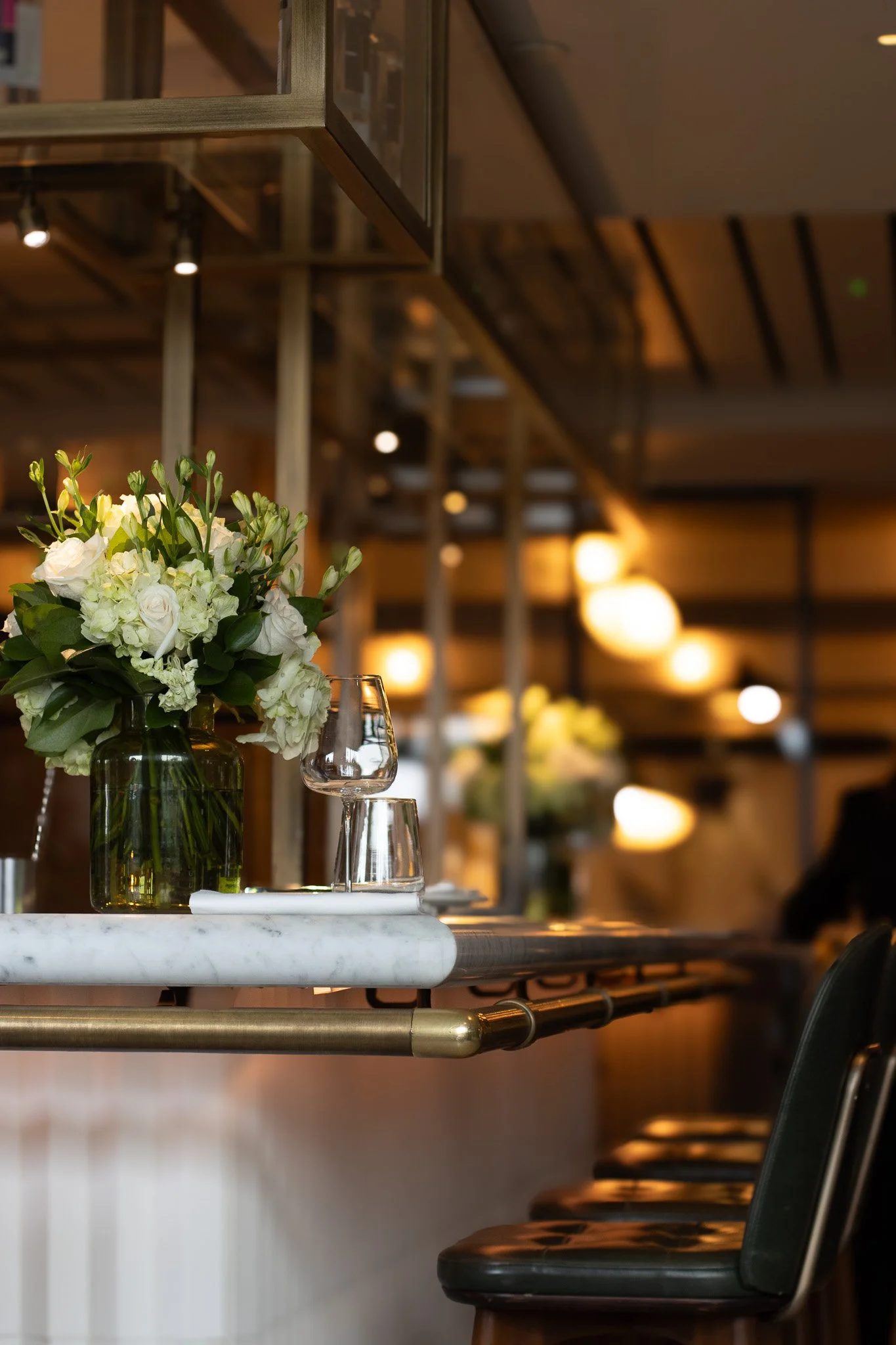 A bar or restaurant interior with a white marble counter, a bouquet of white flowers in a green glass vase, and empty wine glasses. Black chairs are lined up along the counter, and warm lighting creates a cozy atmosphere. The Hari, Il Pampero.