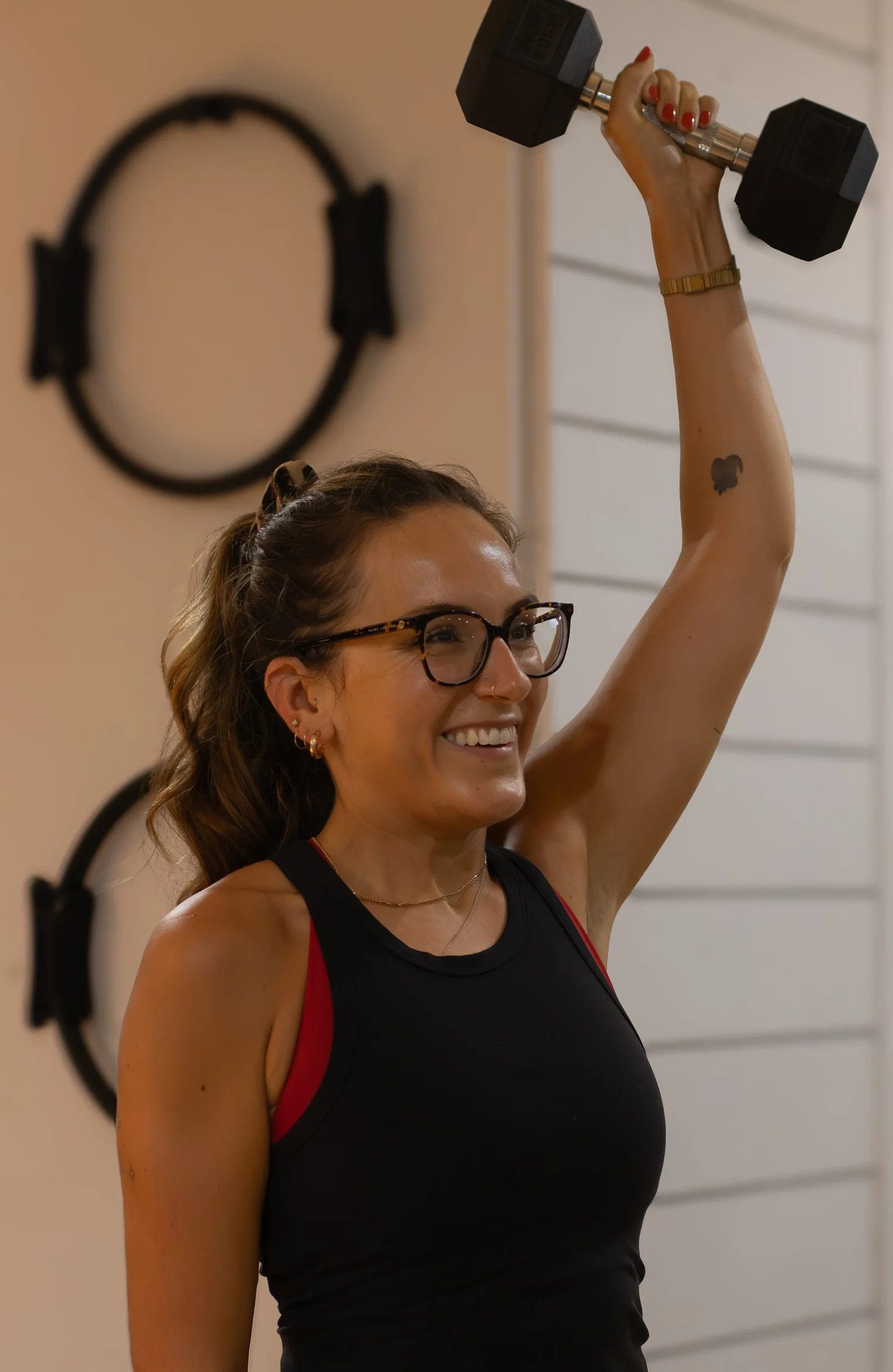A smiling woman wearing glasses and a black and red workout tank top raises a black dumbbell in her right hand during a fitness session in a gym. The Studio Cheltenham, Reformer Pilates.