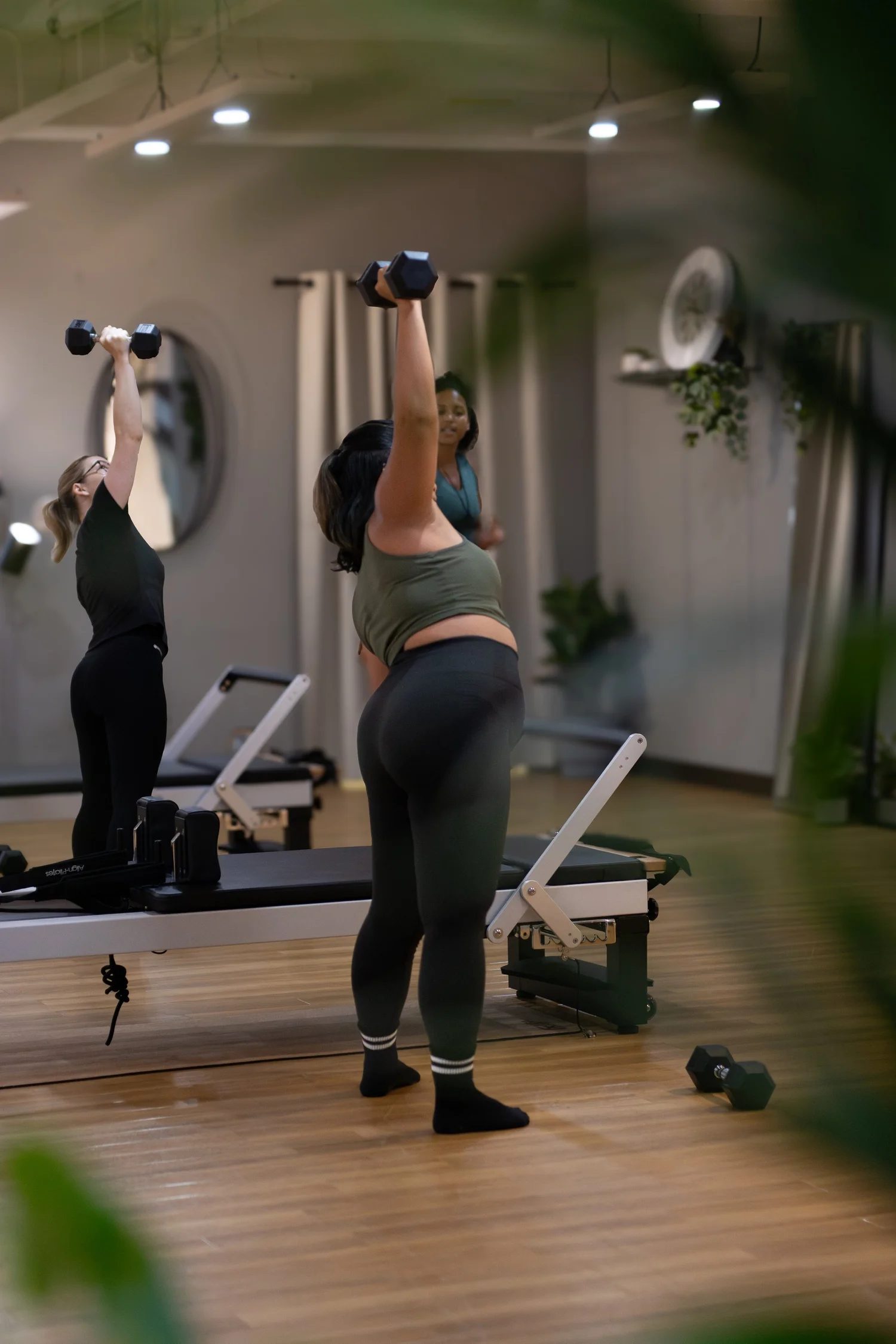 Two women lifting dumbbells during a workout in a gym, with exercise machines and wall decor visible in the background. The Studio Cheltenham, Reformer Pilates.