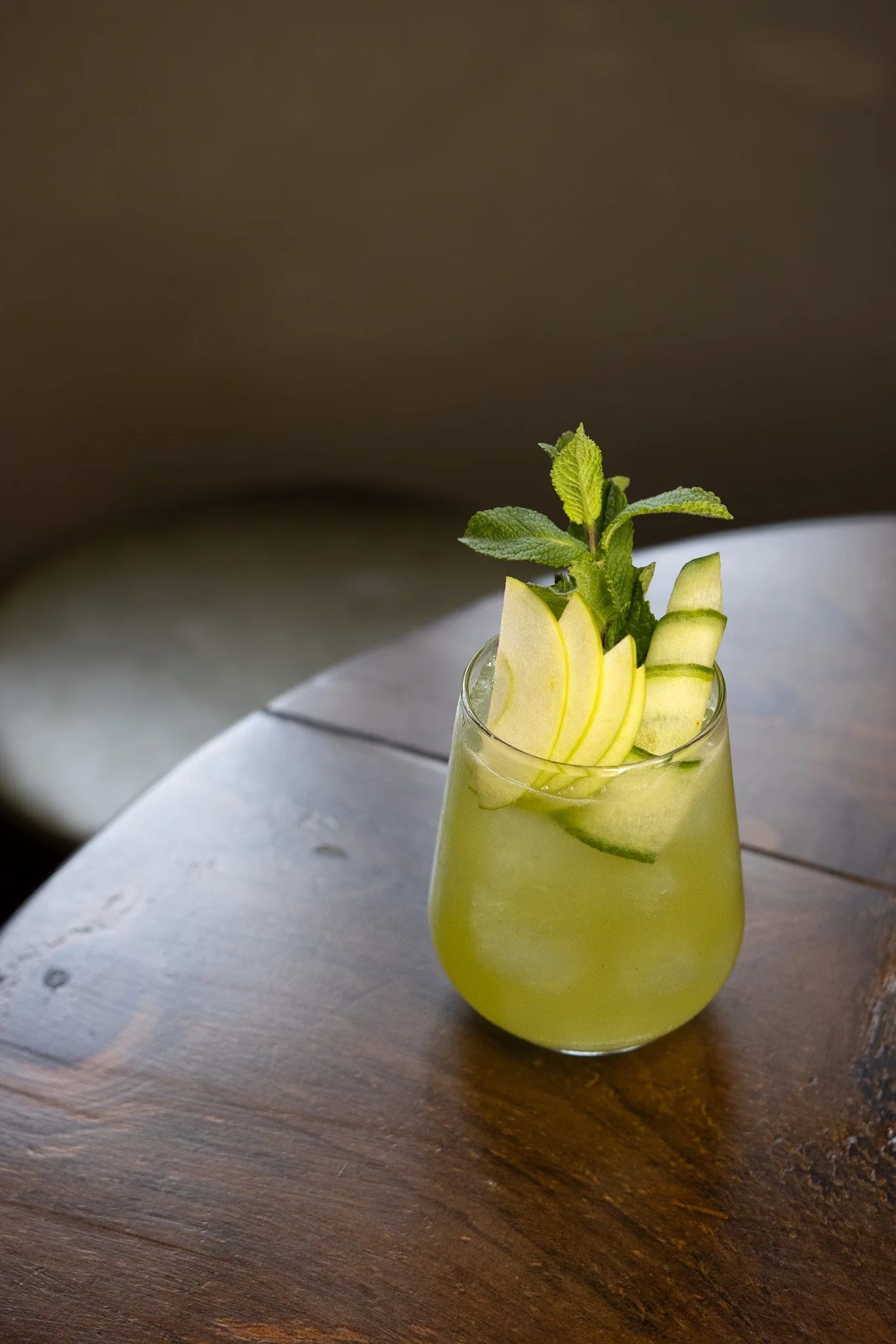 A glass of yellow-green beverage garnished with apple slices and mint leaves, placed on a wooden table. The Lamb Inn. Cocktails. Mixology.
