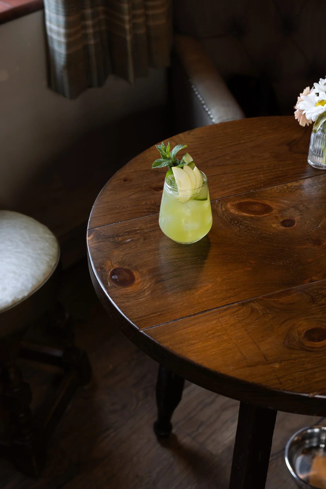 A wooden table with a green drink garnished with apple slices, mint, and a sprig of herbs in a glass, with a floral arrangement in a glass vase nearby. The Lamb Inn.