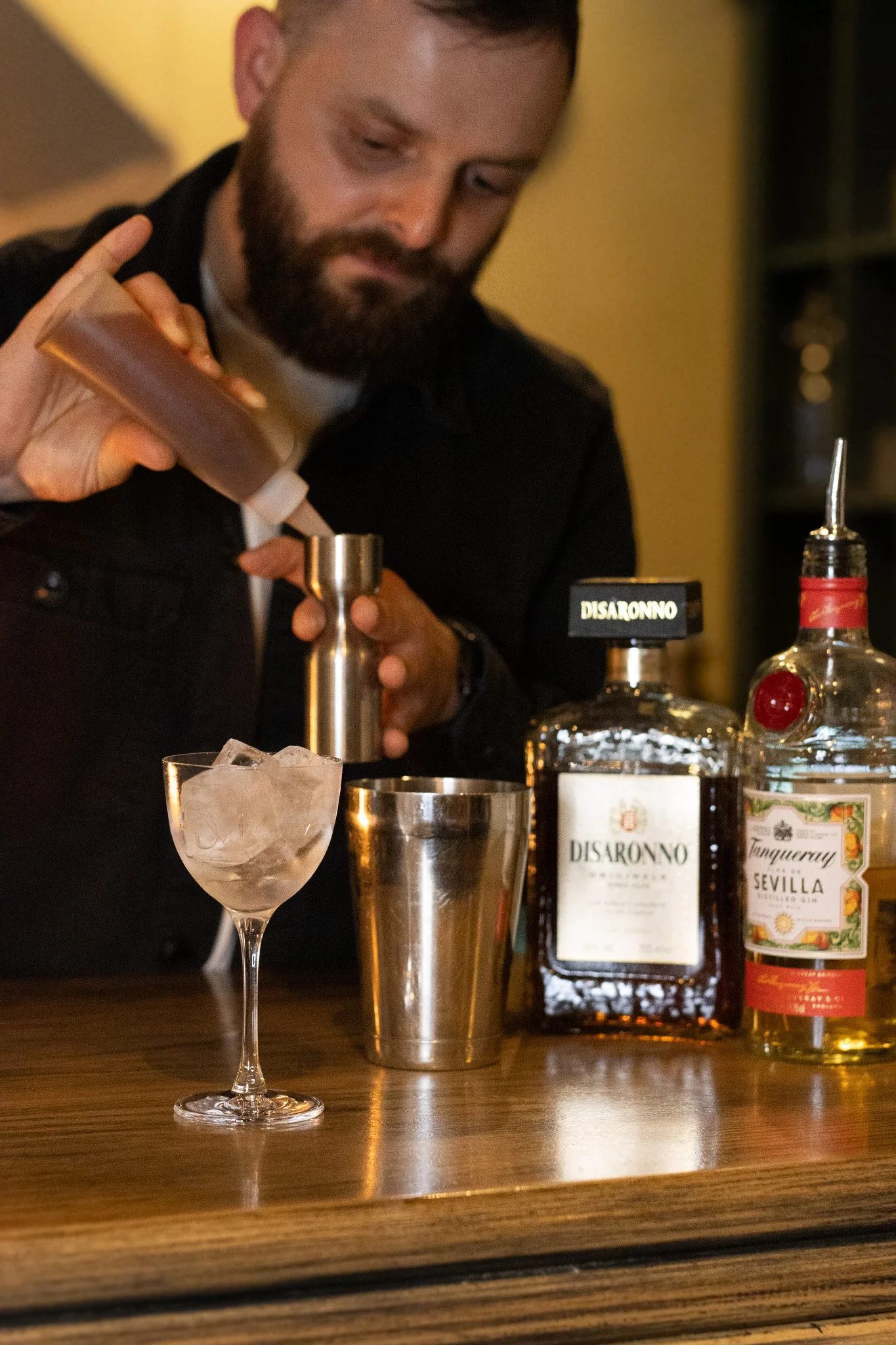 A bartender preparing a cocktail behind a bar. The image shows a glass with ice, bottles of alcohol including Disaronno and Tanqueray gin, and bar tools. The Lamb Inn.