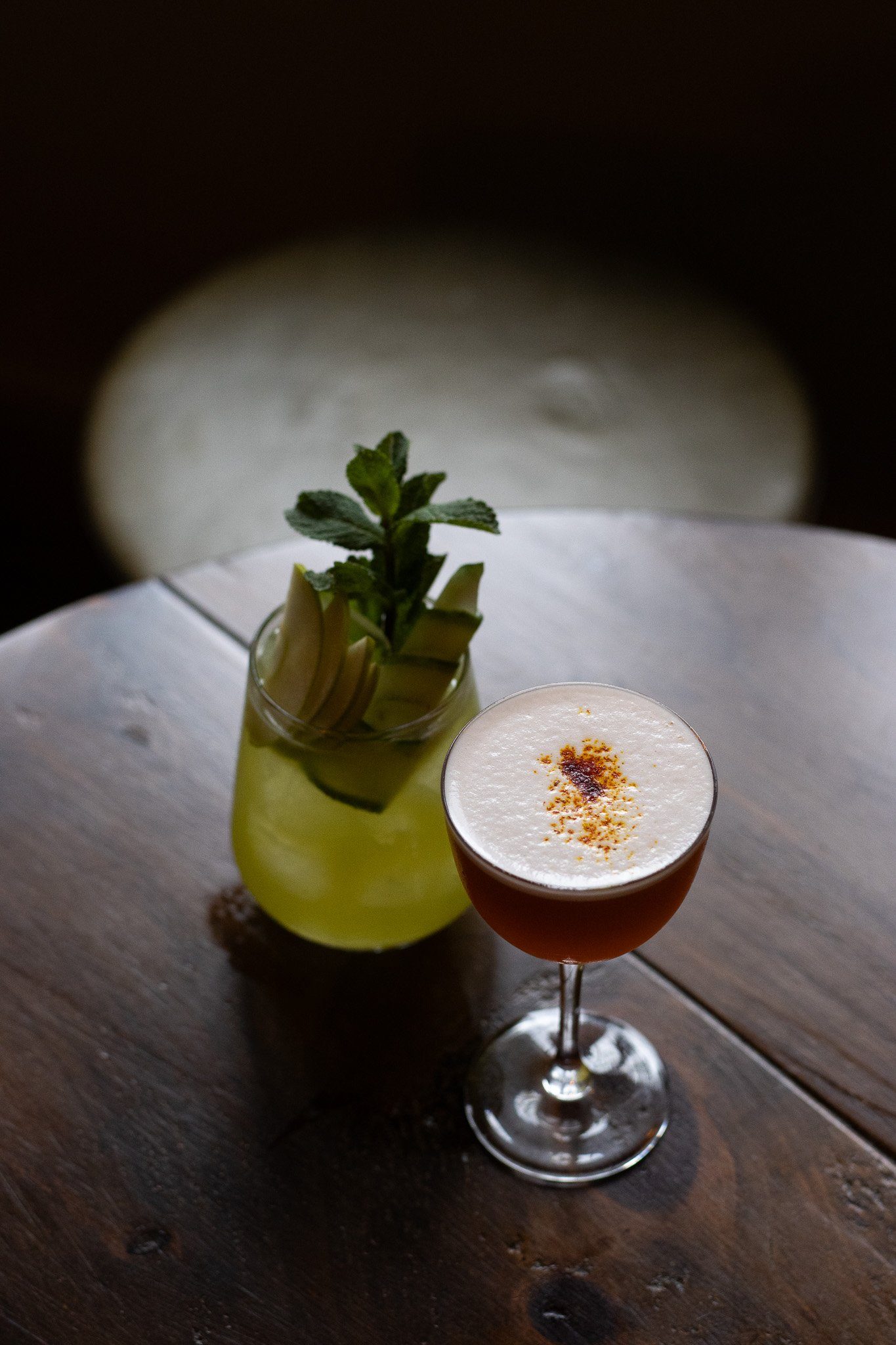 A cocktail with a foamy top garnished with a sprinkle of spice, positioned in a glass on a wooden table with a planter containing a green plant in the background.