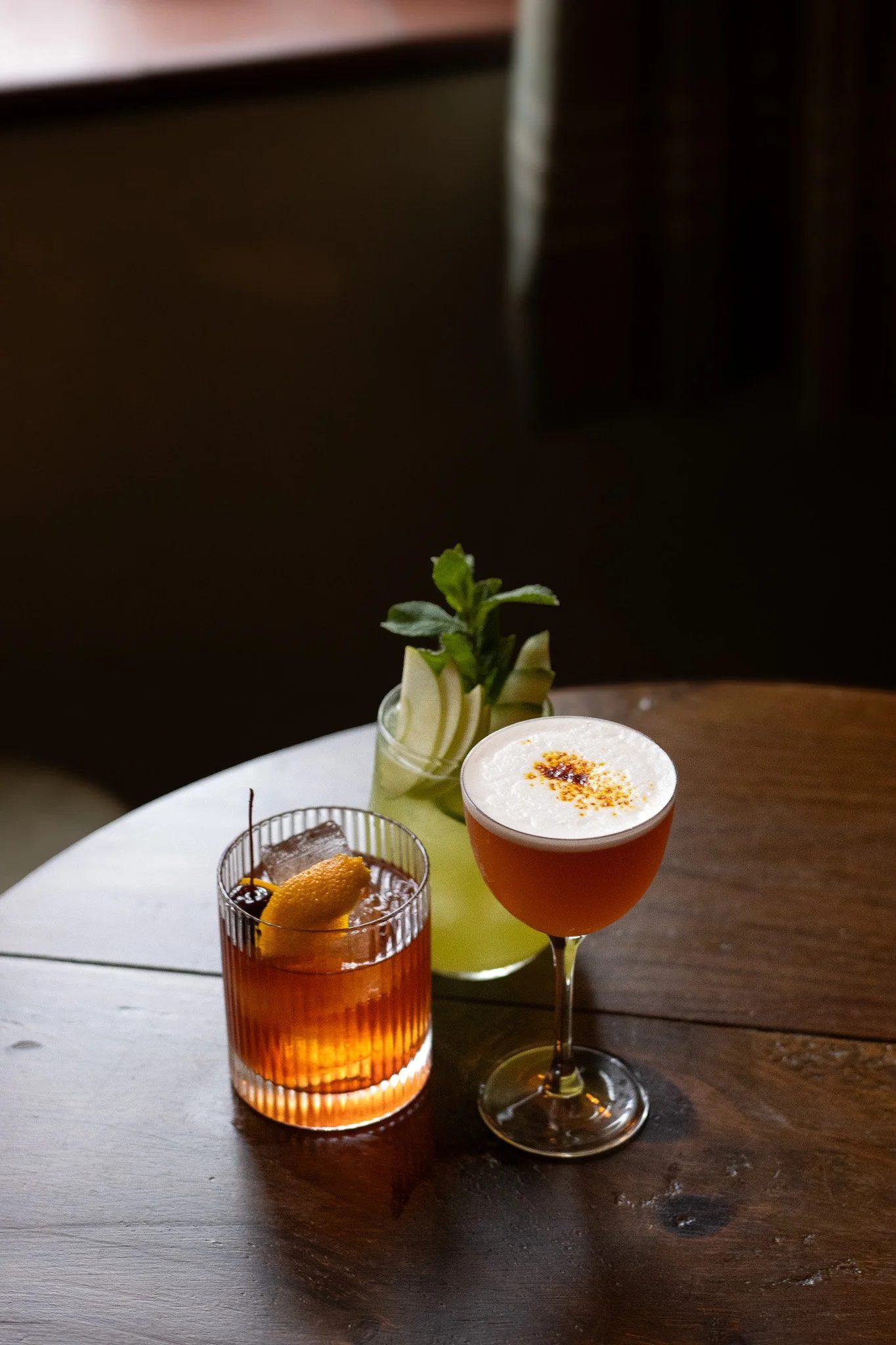 Three cocktails on a round table: a blue drink with orange peel, an amber drink with cherry, and a pink cocktail with frothy top and spices, with a plant in the background. The Lamb Inn.