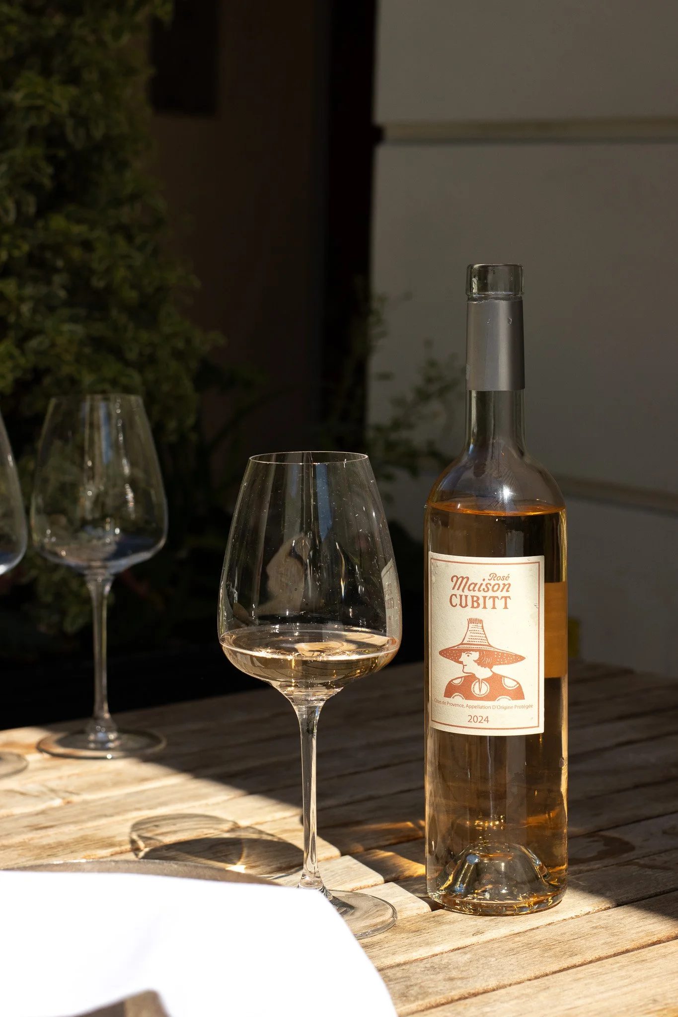 A bottle of rosé wine labeled 'Maison Cubitt' with a drawing of a woman wearing a hat, and three empty wine glasses on a wooden table. Alfred Tennyson.