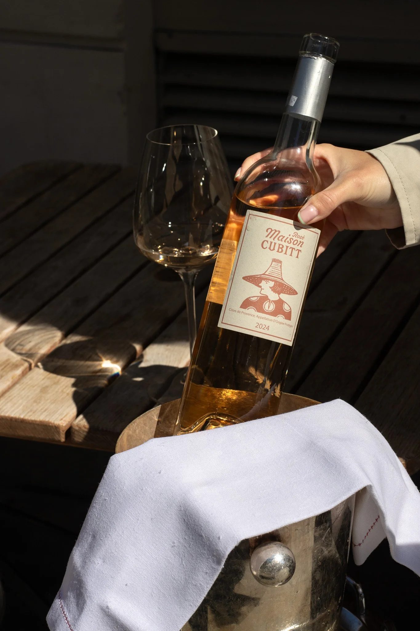 A hand holding a bottle of Maison Cubitt Rosé wine over a metal ice bucket, on a wooden outdoor table with a glass of water nearby, and a white cloth draped over the bucket. Alfred Tennyson.
