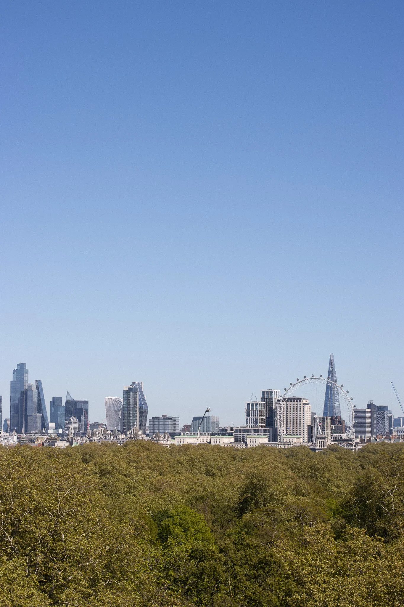 London skyline with trees in the foreground, including the Shard and the London Eye, under a clear blue sky. The Peninsula.
