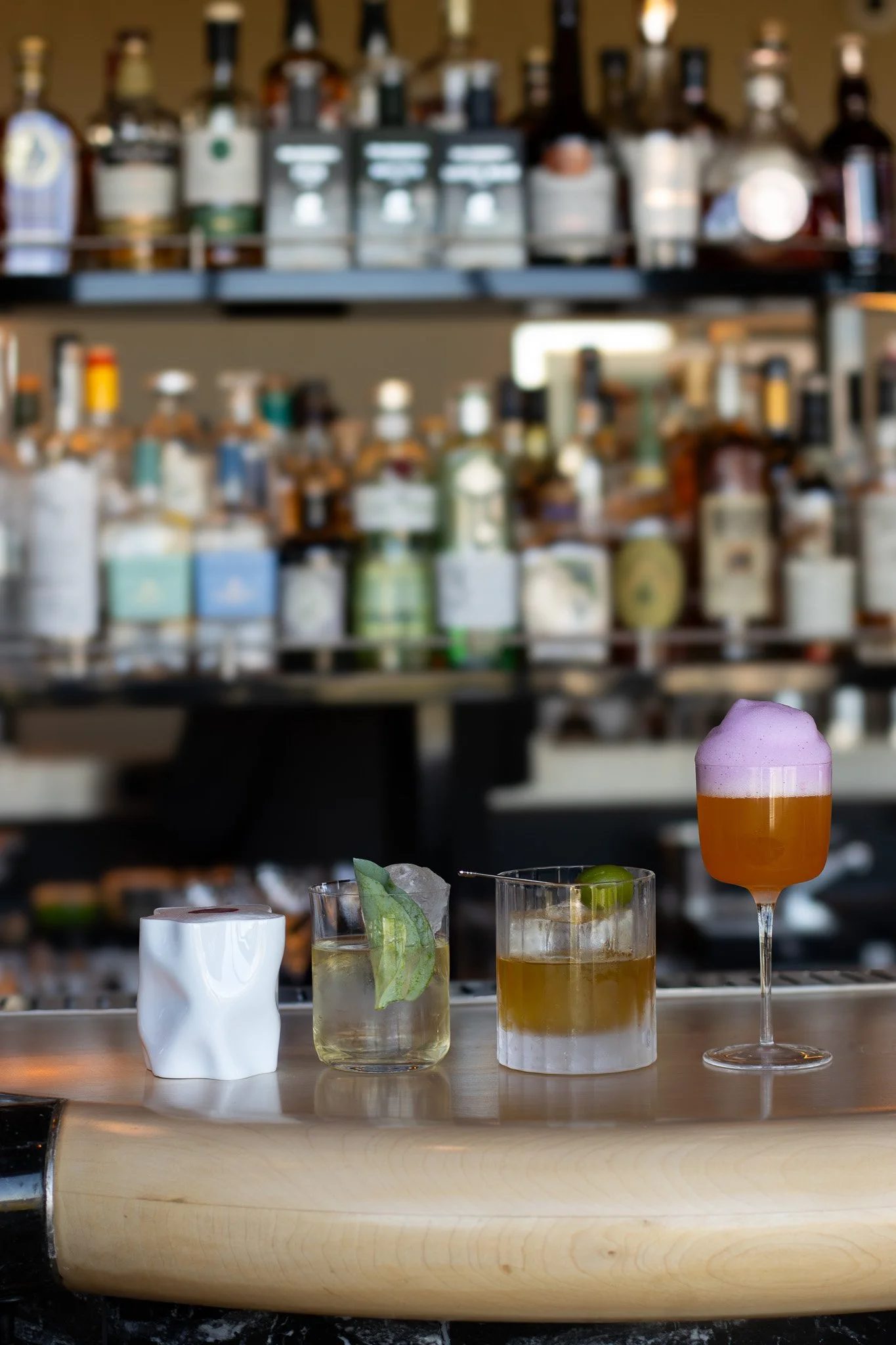 A bartender's countertop with various cocktails and garnishes, including a purple foam-topped drink in a wine glass, a drink with lime and a leaf, and a cocktail with a green garnish, set against a background of shelves filled with liquor bottles. Th