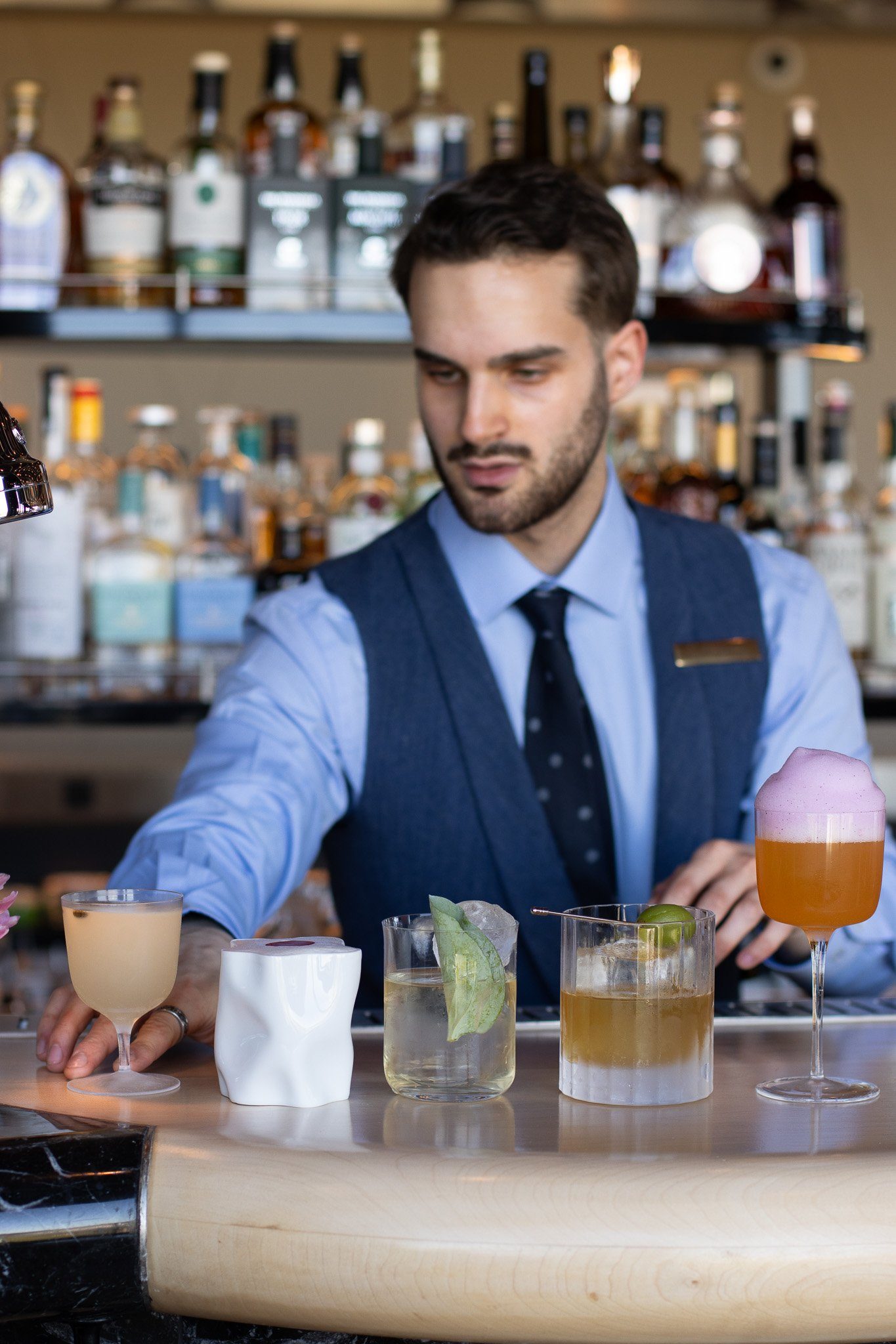 Bartender mixing drinks at a bar with various bottles in the background. Several colorful cocktails are on the bar counter. The Peninsula.