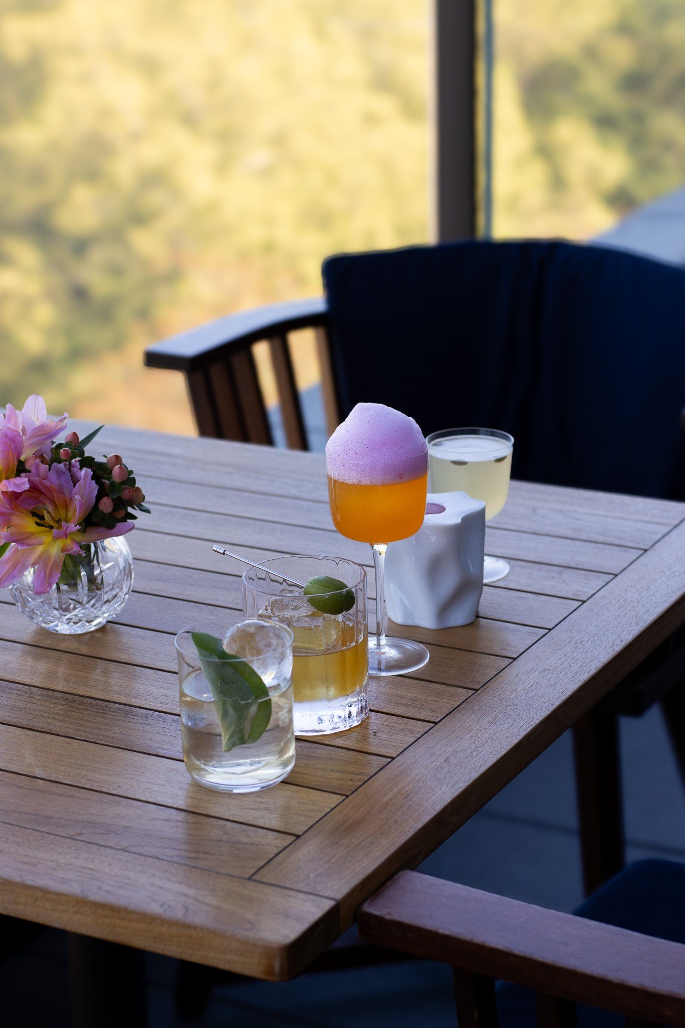 A wooden table set with various drinks, including cocktails with foam and lime, and a small vase of pink flowers. There is a blurred background of a window with green landscape outside. The Peninsula.