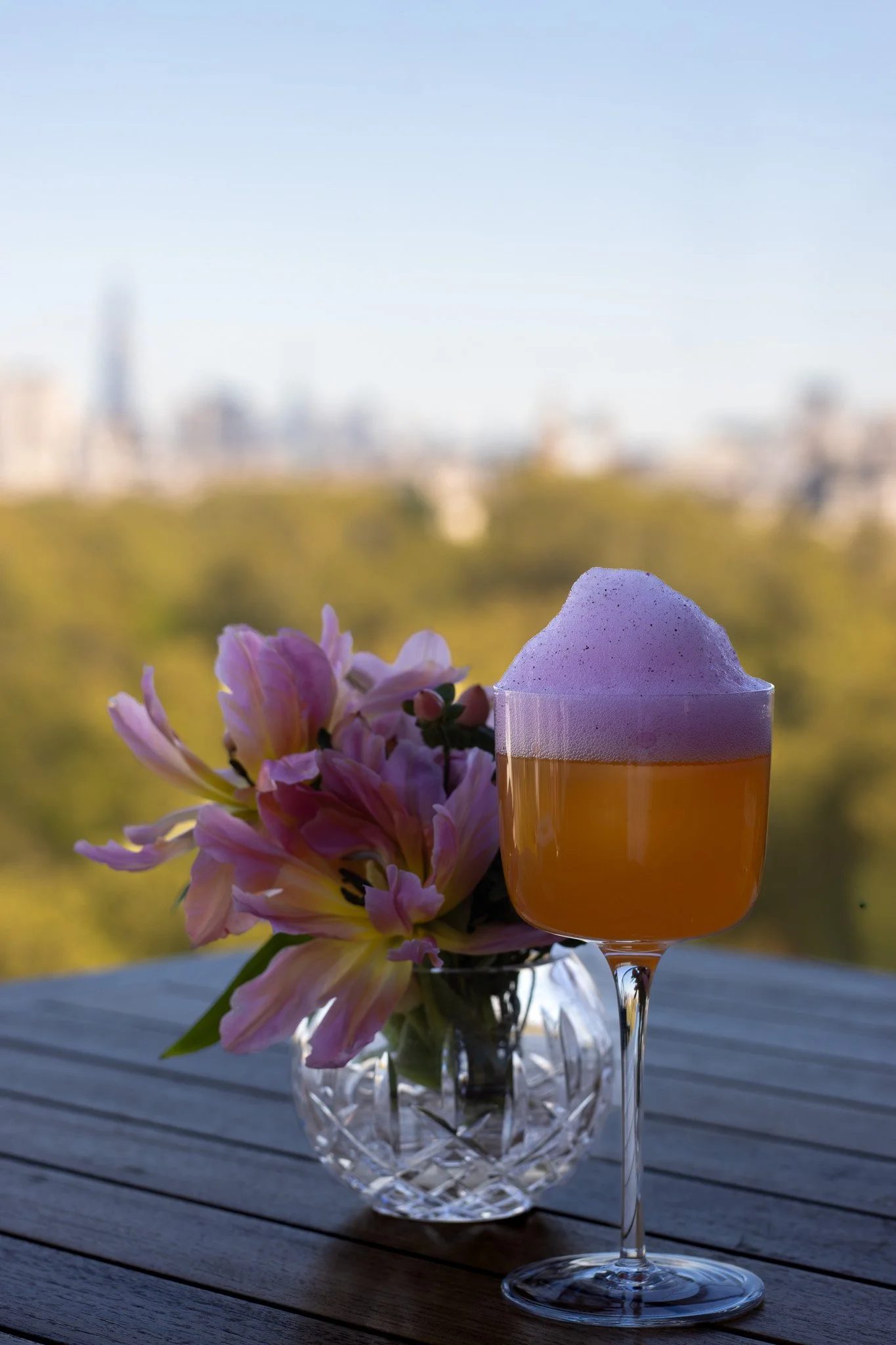 A glass of orange beverage topped with foam, placed on a wooden outdoor table with a bouquet of pink and yellow flowers in a crystal vase, against a blurred cityscape background. The Peninsula.