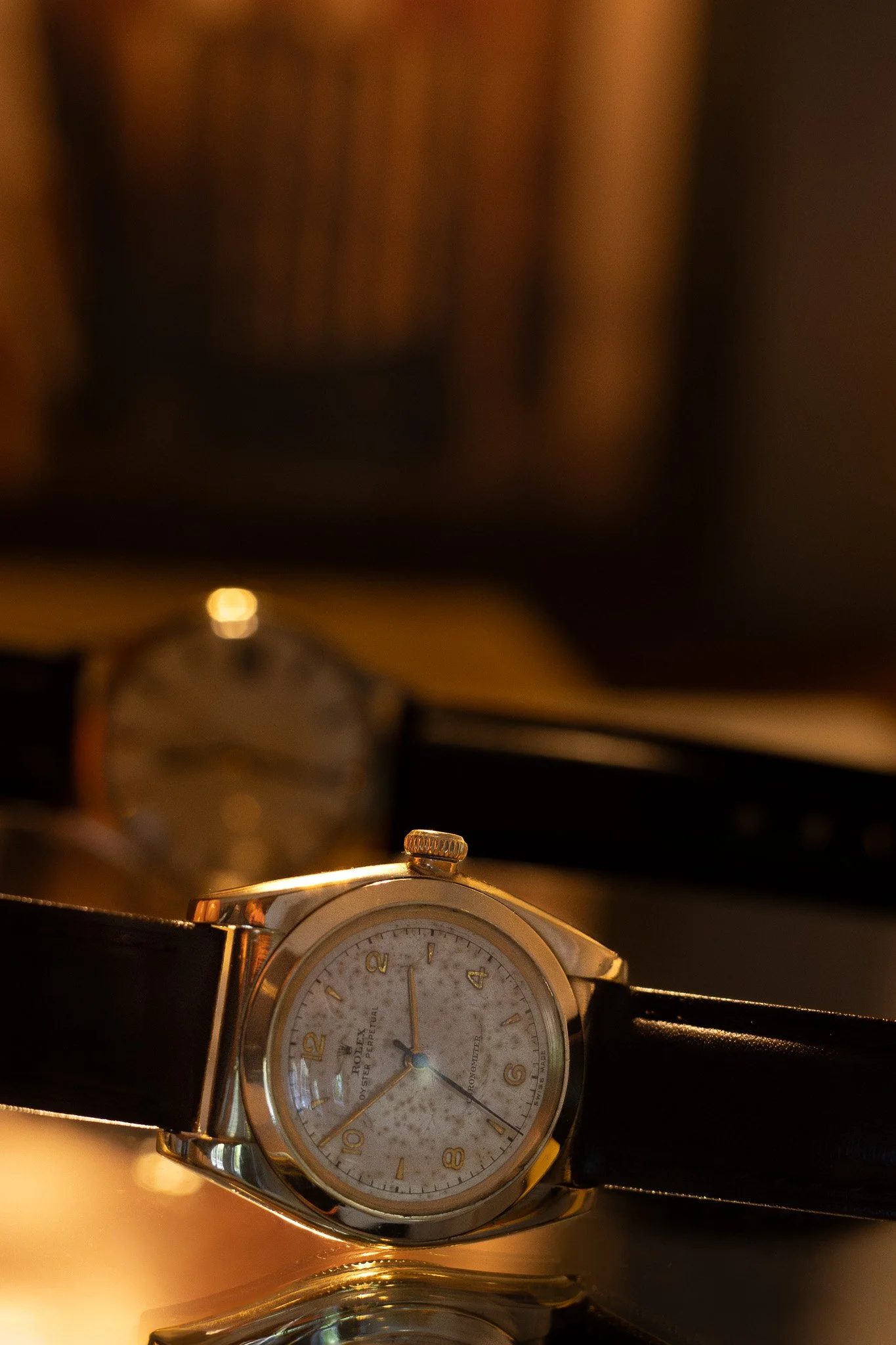 A wristwatch with a white dial, gold case, black leather strap, and gold crown, placed on a shiny surface, with a blurred background of warm ambient lighting. Aurmont. Watches.