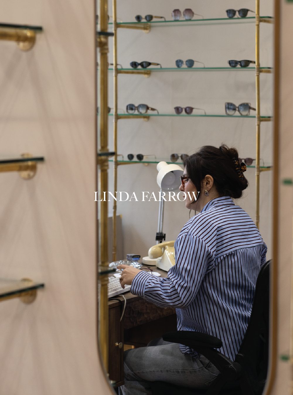 A woman with dark hair in a bun, wearing glasses and a striped shirt, sitting at a desk with a vintage beige rotary telephone, a lamp, and some small items, surrounded by shelves with various sunglasses on display. Linda Farrow.